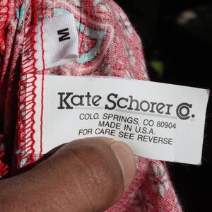 May include: A close-up of a clothing tag with the text "Kate Schorer C.", "COLO. SPRINGS, CO 80904", and "MADE IN U.S.A.". The tag is white with black text and a small "M" tag. The fabric is red with a white and teal pattern.