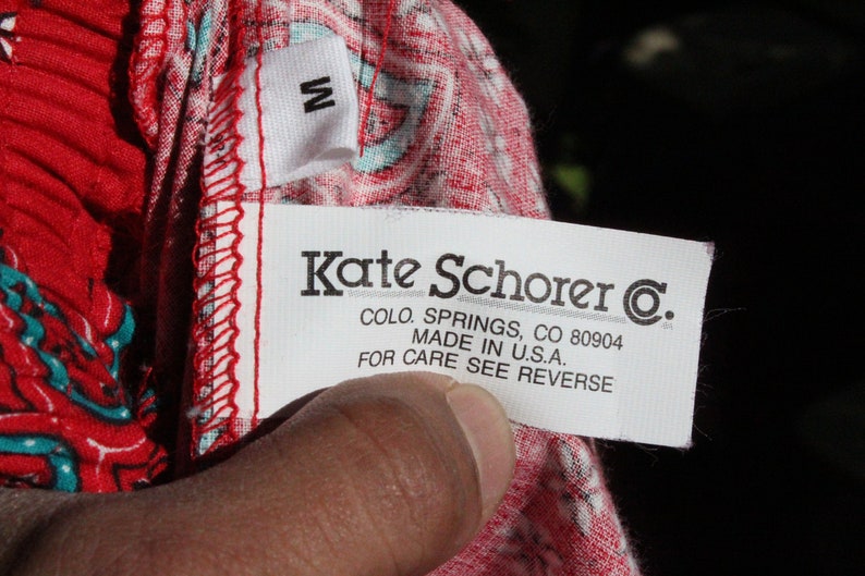 May include: A close-up of a clothing tag with the text "Kate Schorer C.", "COLO. SPRINGS, CO 80904", and "MADE IN U.S.A.". The tag is white with black text and a small "M" tag. The fabric is red with a white and teal pattern.