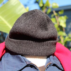 May include: A dark gray knit beanie hat. The hat is made of a textured material and is worn over a blue jacket and a red coat. The background features green leaves and sunlight.