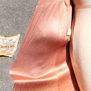 May include: Close-up of a peach-colored satin fabric draped over a mannequin. The fabric has a subtle sheen and is contrasted by a gray wool fabric. A small tag with text is visible.
