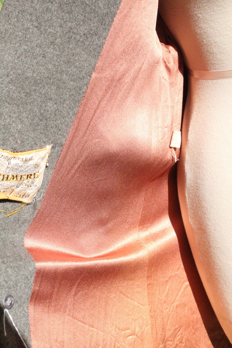 May include: Close-up of a peach-colored satin fabric draped over a mannequin. The fabric has a subtle sheen and is contrasted by a gray wool fabric. A small tag with text is visible.