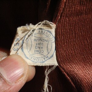 May include: Close-up of a vintage brown fabric with a textured surface. A small, torn tag is attached, featuring a blue circular stamp with text that reads "NATIONAL COAT AND SUIT INDUSTRY".
