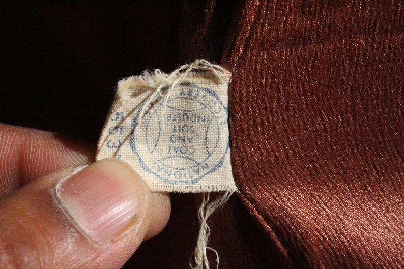 May include: Close-up of a vintage brown fabric with a textured surface. A small, torn tag is attached, featuring a blue circular stamp with text that reads "NATIONAL COAT AND SUIT INDUSTRY".