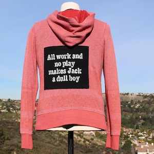 May include: A red hoodie with a black square patch on the back. The patch features white text that reads "All work and no play makes Jack a dull boy." The hoodie has a drawstring hood and ribbed cuffs.