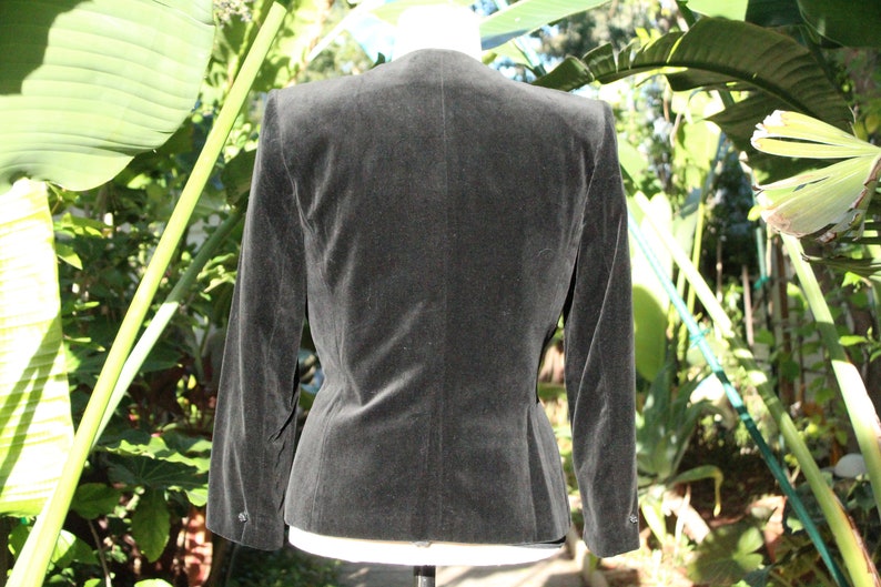 May include: A dark gray velvet blazer with a tailored fit, featuring a classic collar and buttoned cuffs. The jacket is displayed on a mannequin, showcasing its structured silhouette and elegant design. The blazer is a versatile piece for various occasions.