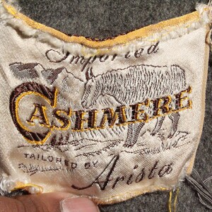 May include: Close-up of a fabric label with the word "CASHMERE" embroidered in gold thread. The label also features an embroidered illustration of an animal and the words "Imported" and "Tailored by Arista". The label is on a gray fabric.