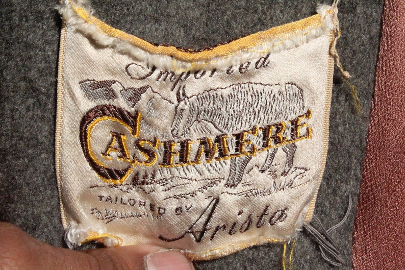 May include: Close-up of a fabric label with the word "CASHMERE" embroidered in gold thread. The label also features an embroidered illustration of an animal and the words "Imported" and "Tailored by Arista". The label is on a gray fabric.