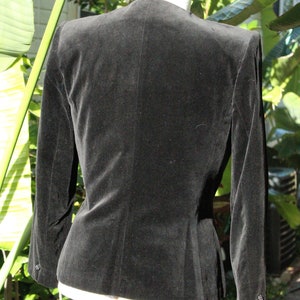 May include: A black velvet blazer with a tailored fit, featuring long sleeves and a classic collarless design. The back view showcases the jacket's smooth texture and subtle shaping, perfect for formal or semi-formal wear.