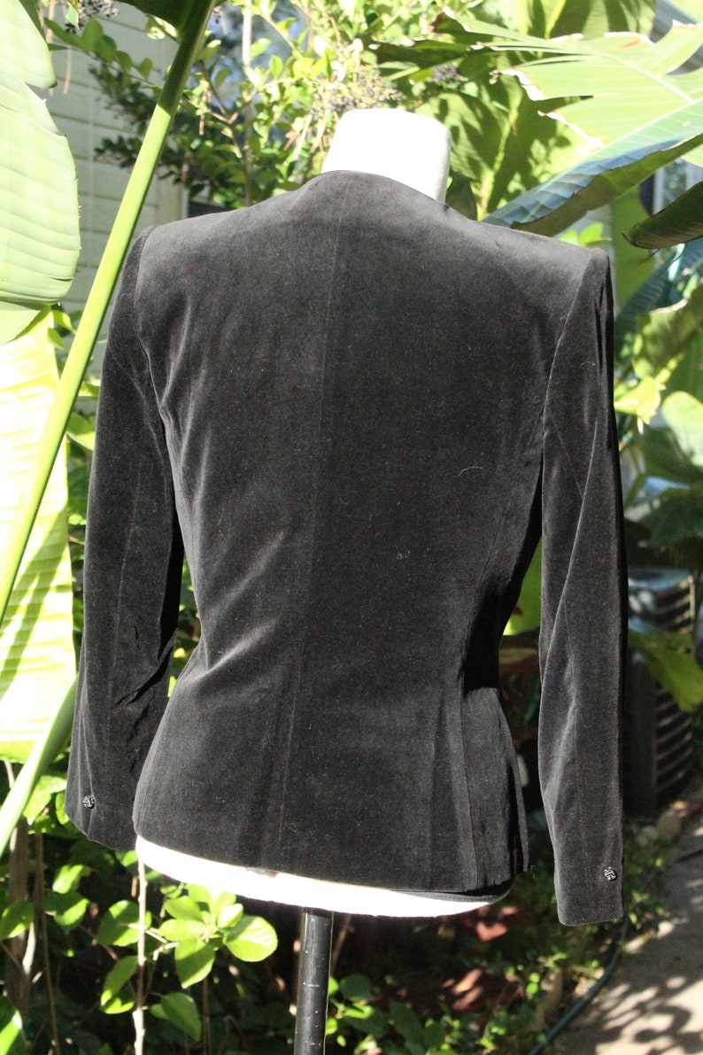 May include: A black velvet blazer with a tailored fit, featuring long sleeves and a classic collarless design. The back view showcases the jacket's smooth texture and subtle shaping, perfect for formal or semi-formal wear.