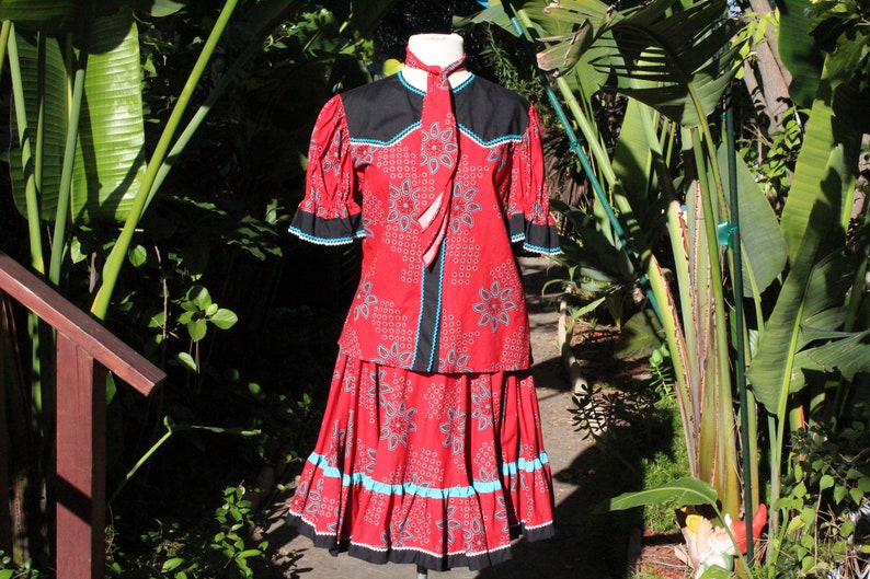 May include: A red two-piece outfit with a floral pattern, black accents, and turquoise trim. The top has short sleeves and a matching tie. The skirt is a similar length. The outfit is displayed outdoors.