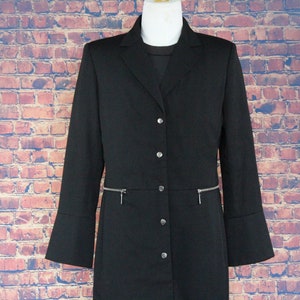 May include: A black long coat with a notched collar, button closure, and two zipped pockets. The coat has long sleeves with flared cuffs. The coat is displayed on a mannequin against a brick wall background.