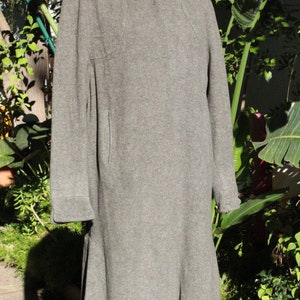 May include: A gray wool coat with a rounded collar and long sleeves. The coat has a button closure and a flared skirt. The coat is displayed on a mannequin in front of a green leafy background.