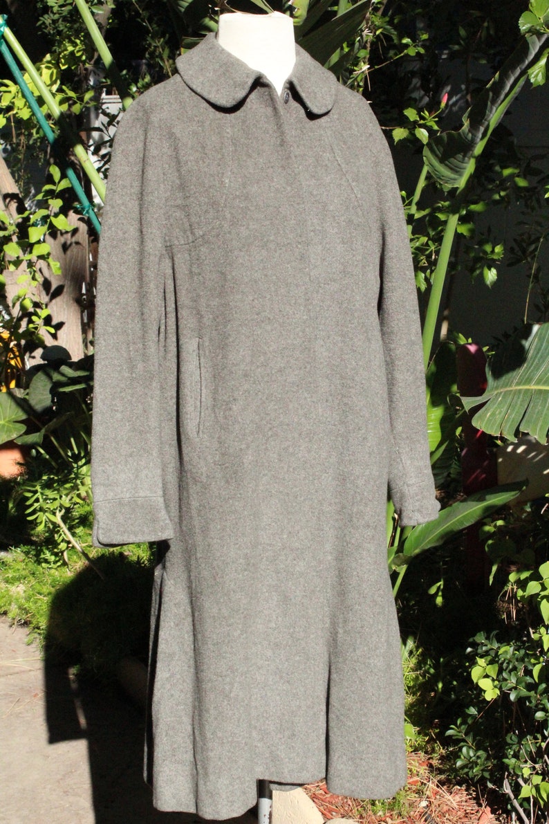 May include: A gray wool coat with a rounded collar and long sleeves. The coat has a button closure and a flared skirt. The coat is displayed on a mannequin in front of a green leafy background.