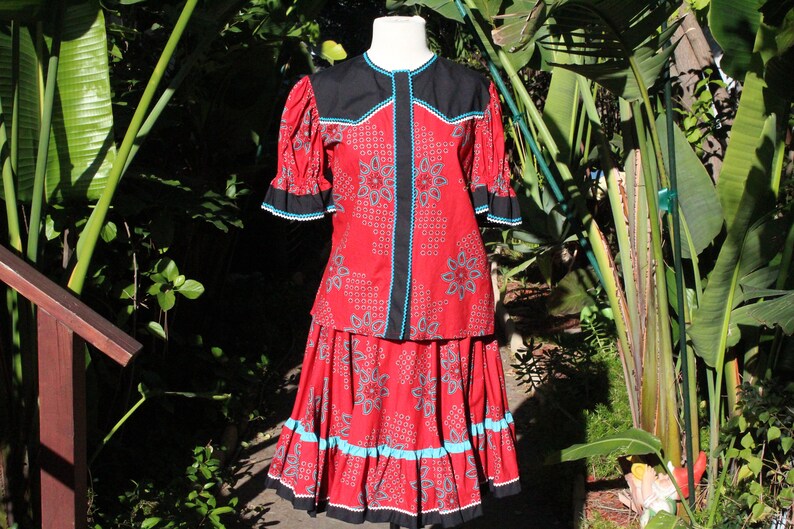 May include: A red two-piece outfit with a floral pattern, black accents, and turquoise trim. The top has short sleeves and a round neckline. The skirt is a matching length. The outfit is displayed on a mannequin.