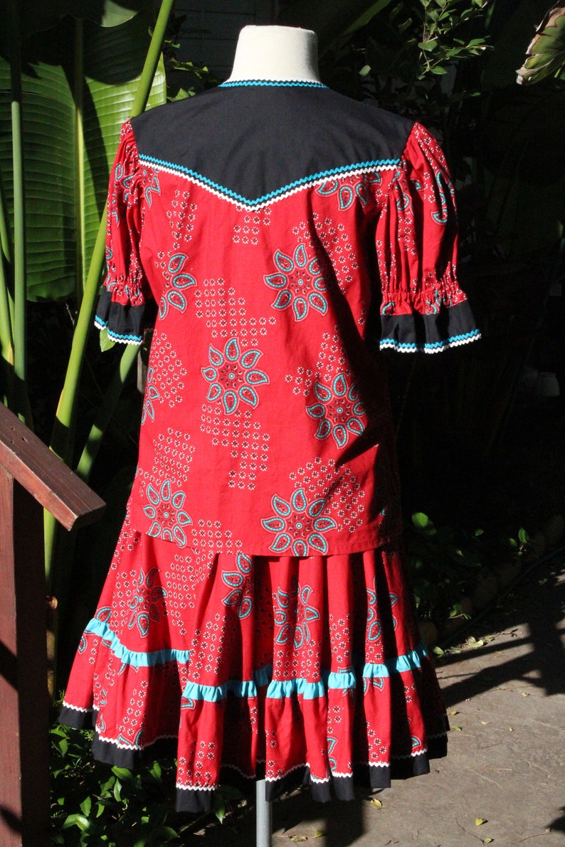 May include: A red two-piece outfit with a floral pattern and turquoise accents. The top has a black yoke and ruffled sleeves. The skirt has a tiered design with turquoise and black trim. The outfit is displayed on a mannequin.