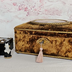 Può includere: Una collezione di oggetti decorativi antichi, tra cui un grande portagioie in velluto marrone con finiture dorate e una chiave con nappa rosa. Altri oggetti includono una piccola scatola nera, un busto e un supporto decorativo.