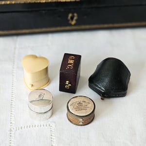 May include: A collection of vintage jewellery boxes and containers. Features a heart-shaped ivory box, a small rectangular box with "G.M.C.", a black leather box, a silver container with "B", and a round container with text.