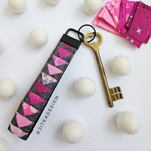 May include: A pink and gray fabric key chain with a black metal ring and key loop. The key chain is made of small fabric triangles sewn together. The key chain is surrounded by white pom-poms.