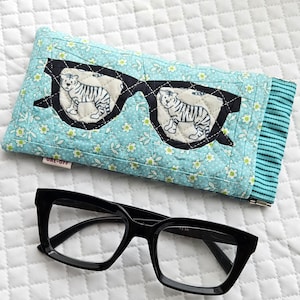 Könnte beinhalten: Ein gestepptes Brillenetui mit hellblauem Blumenmuster und einer schwarzen Umrandung einer Sonnenbrille, wobei jedes Brillenglas eine weiße Tigerillustration zeigt. Daneben eine schwarze Brille mit quadratischem Rahmen.