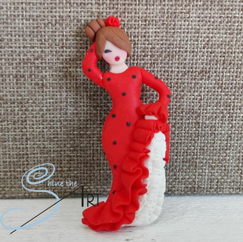 May include: A red and white figurine of a woman in a traditional Spanish dress with black polka dots. The figurine is made of polymer clay and has a white ruffle on the skirt.