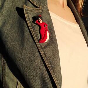 May include: A red and white flamenco dancer figurine with black polka dots, worn as a pin on a denim jacket.