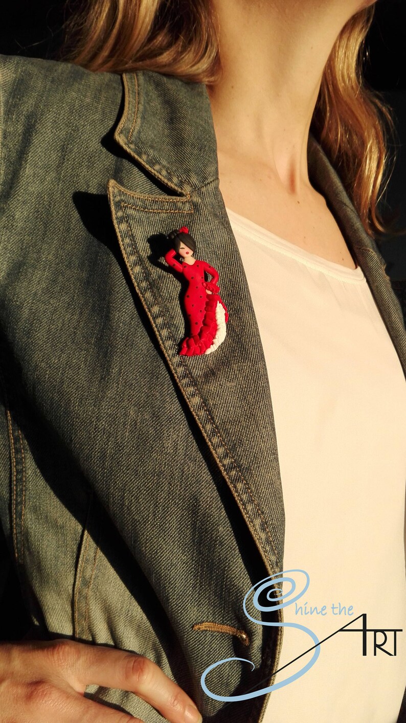 May include: A red and white flamenco dancer figurine with black polka dots, worn as a pin on a denim jacket.