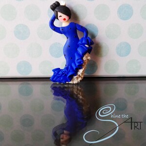 May include: A blue and white figurine of a flamenco dancer with a polka dot dress and a white ruffle skirt. The figurine is standing on a reflective surface with a polka dot pattern.