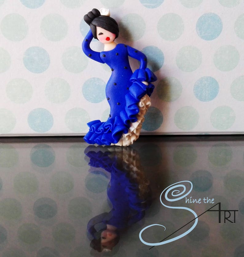 May include: A blue and white figurine of a flamenco dancer with a polka dot dress and a white ruffle skirt. The figurine is standing on a reflective surface with a polka dot pattern.