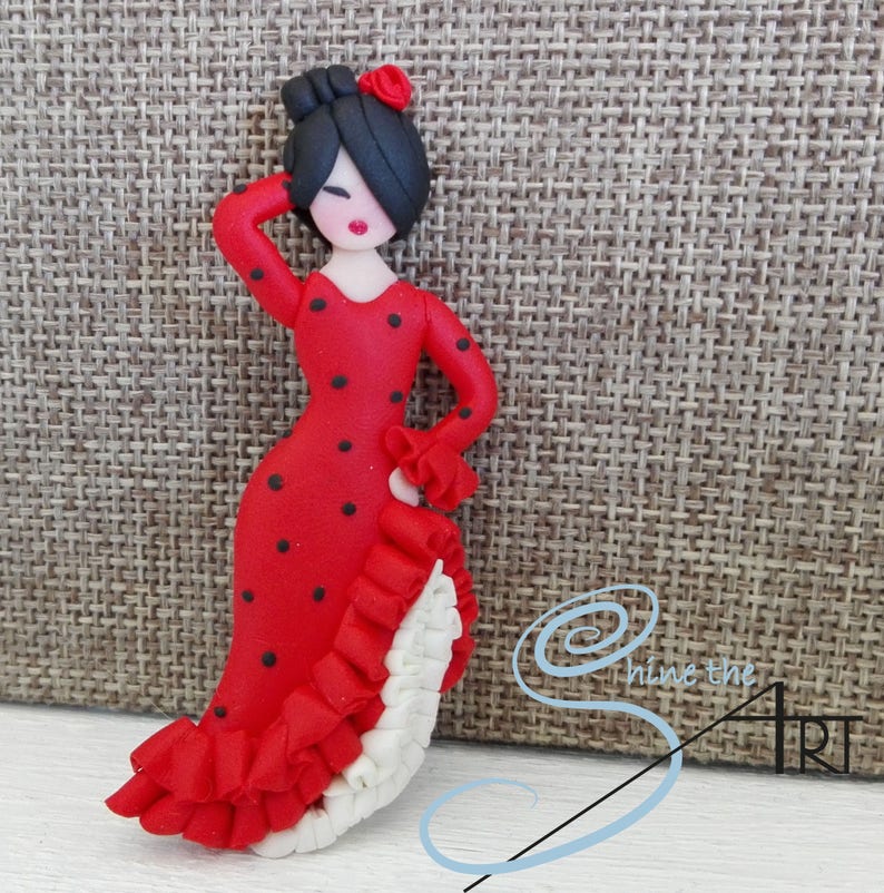 May include: A red and black polka dot dress with white ruffles. The dress is worn by a small doll with black hair and a red flower.