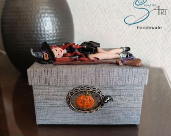 Könnte beinhalten: Eine dekorative graue Box mit einem Kürbis-Emblem, gekrönt von einer Miniatur-Hexenfigur und einem Besen. Die Hexe trägt einen schwarzen Hut und ein schwarzes Kleid mit roten Haaren. Auf der Box steht "shine the art handmade".