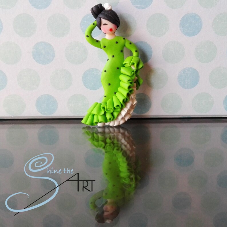 May include: A green and black polka dot dress with a white ruffle trim. The dress is worn by a small figurine of a woman with black hair and red lips. The figurine is standing on a reflective surface.