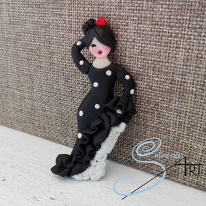 May include: A black and white polka dot flamenco dancer figurine with a red flower in her hair. The figurine is made of polymer clay and is standing on a white base.