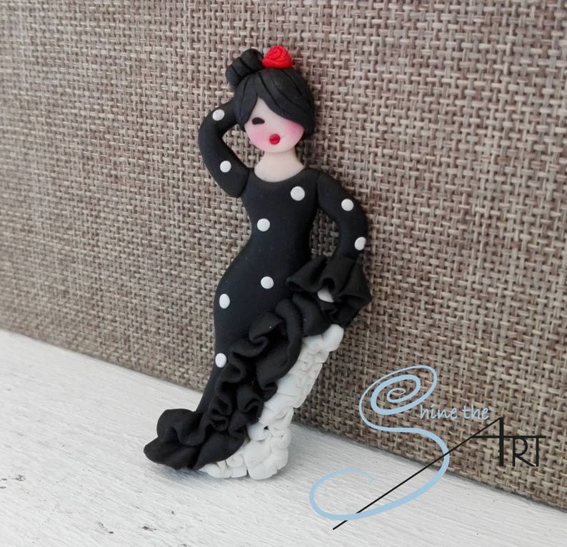 May include: A black and white polka dot flamenco dancer figurine with a red flower in her hair. The figurine is made of polymer clay and is standing on a white base.