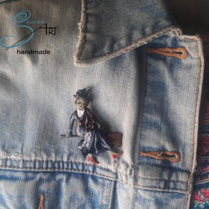 Puede incluir: Una figura de bruja hecha a mano sobre una escoba, sujeta a una chaqueta vaquera. La bruja lleva un sombrero negro, un vestido azul oscuro con detalles plateados y zapatos rojos. La chaqueta tiene costuras y botones marrones. El texto "shine the art handmade" es visible.