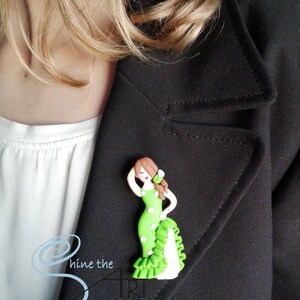 May include: A green and white polka dot dress with a white ruffle and a brown haired woman wearing it. The woman is holding her hand to her head and has a flower in her hair. The dress is a handmade polymer clay brooch.