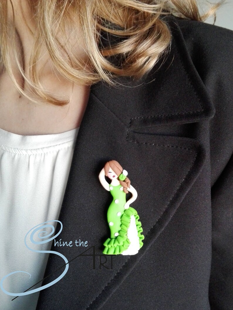 May include: A green and white polka dot dress with a white ruffle and a brown haired woman wearing it. The woman is holding her hand to her head and has a flower in her hair. The dress is a handmade polymer clay brooch.