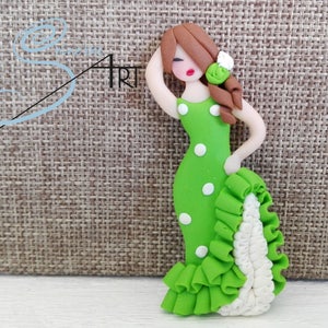 May include: A green and white polka dot dress with a white ruffle trim. The dress is made of polymer clay and is a figurine of a woman with brown hair and red lips.