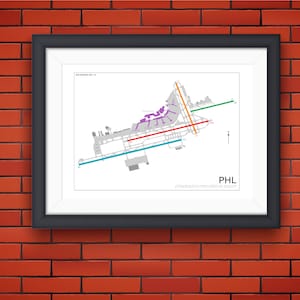 May include: A black and white diagram of Philadelphia International Airport (PHL) showing runways, taxiways, and terminal buildings. The diagram is framed in a black frame and hanging on a red brick wall.