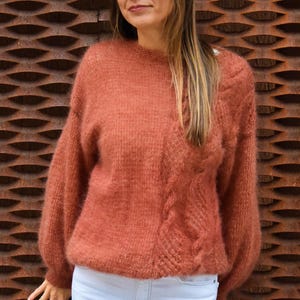 Puede incluir: Un suéter de color óxido con un diseño de punto trenzado en la parte delantera se combina con jeans blancos y un sombrero blanco. El suéter tiene un aspecto suave y texturizado y mangas largas. El fondo es una pared de metal marrón texturizado.