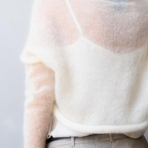 May include: Off-white, sheer knit sweater with long sleeves and a high neckline. The sweater is paired with a white top and light gray plaid pants. The knitwear has a delicate, airy appearance.