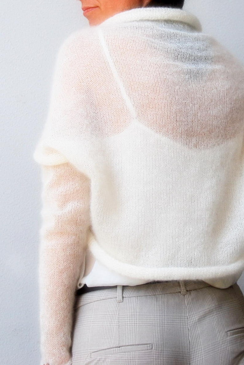 May include: Off-white, sheer knit sweater with long sleeves and a high neckline. The sweater is paired with a white top and light gray plaid pants. The knitwear has a delicate, airy appearance.