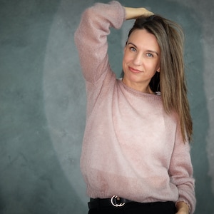 Può includere: Una donna indossa un maglione rosa chiaro a maniche lunghe e una cintura nera. È in piedi davanti a un muro grigio con una texture.