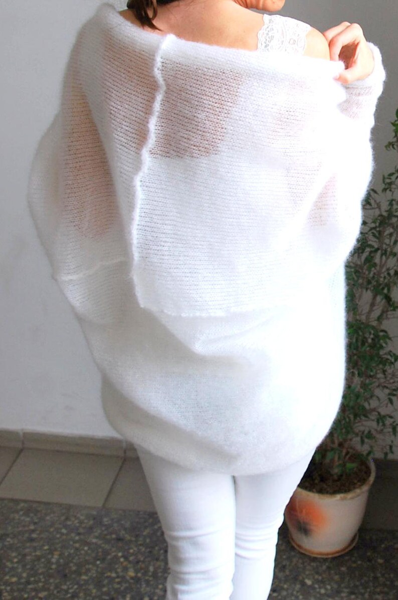 May include: A white, oversized, open-weave sweater. The sweater has a loose, flowing design and is paired with white pants. The image shows the back of the sweater, highlighting its texture and drape. The sweater appears soft and comfortable.