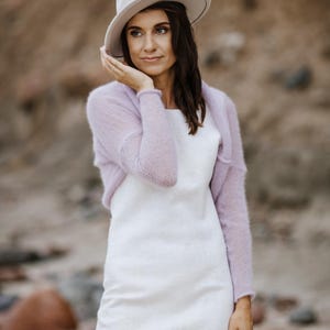 May include: A woman wearing a white dress, a light purple shrug, and a cream-colored hat. The outfit is set against a blurred outdoor background, suggesting a beach or natural setting. The dress is simple, and the shrug adds a touch of color.