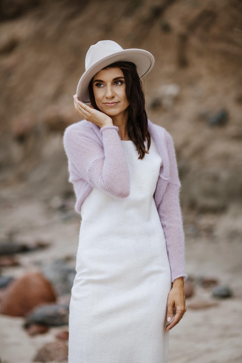May include: A woman wearing a white dress, a light purple shrug, and a cream-colored hat. The outfit is set against a blurred outdoor background, suggesting a beach or natural setting. The dress is simple, and the shrug adds a touch of color.