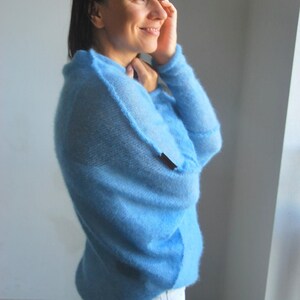 Könnte beinhalten: Eine Frau trägt einen hellblauen, flauschigen Pullover und eine weiße Jeans. Sie steht in der Nähe eines Fensters und schaut zur Seite.