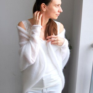 May include: A woman wearing a white outfit, including a lace-trimmed camisole, a sheer, open-front cardigan, and white jeans. The cardigan has long sleeves. The outfit is all white.