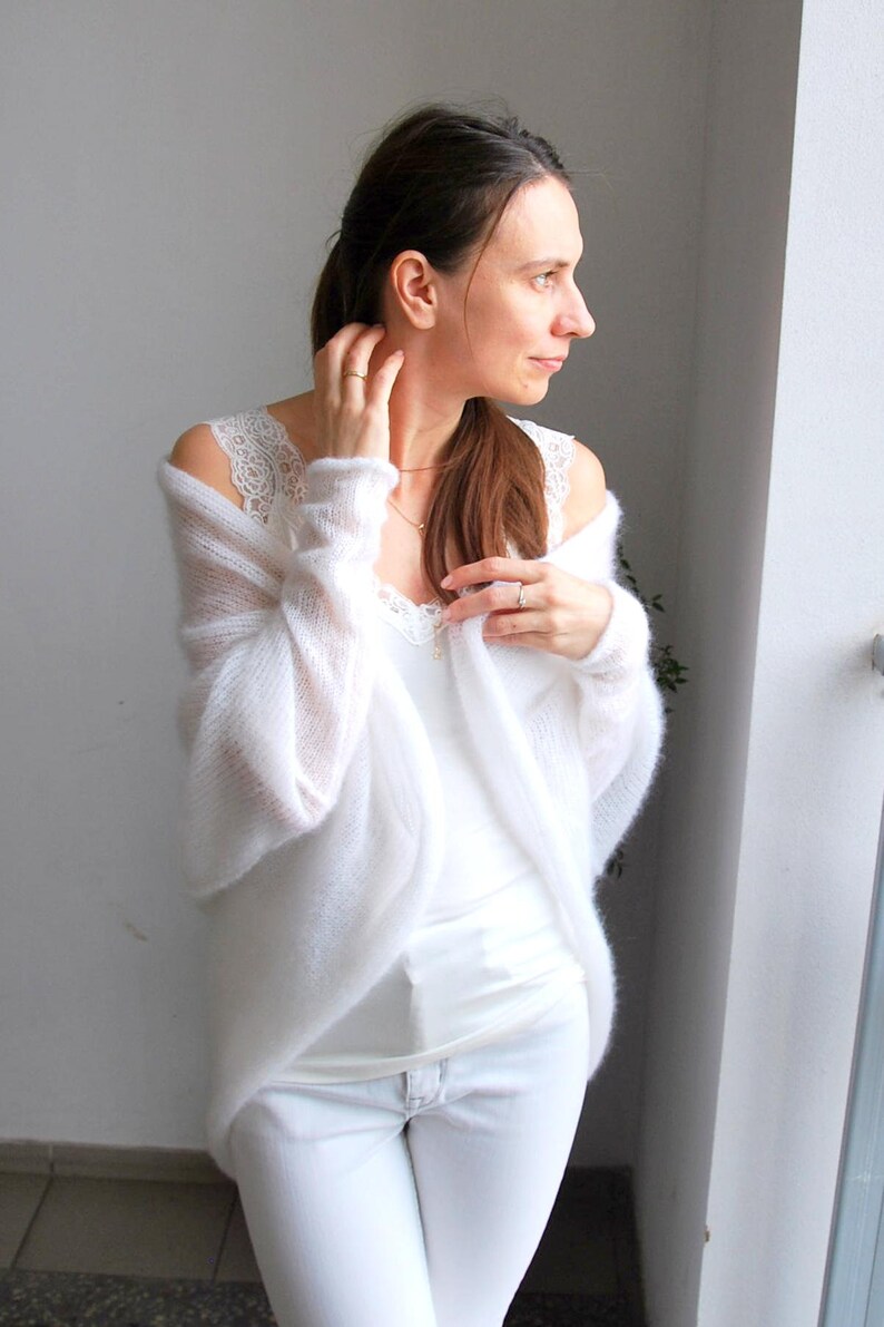 May include: A woman wearing a white outfit, including a lace-trimmed camisole, a sheer, open-front cardigan, and white jeans. The cardigan has long sleeves. The outfit is all white.