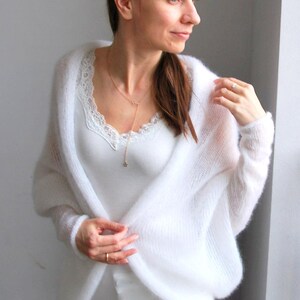 May include: A white, open-front cardigan with long sleeves. The soft, fluffy knitwear is worn over a white lace-trimmed camisole and white pants. A delicate gold necklace with a star pendant adds a touch of elegance to the outfit.