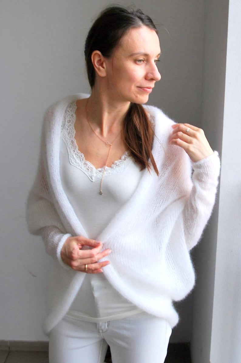 May include: A white, open-front cardigan with long sleeves. The soft, fluffy knitwear is worn over a white lace-trimmed camisole and white pants. A delicate gold necklace with a star pendant adds a touch of elegance to the outfit.